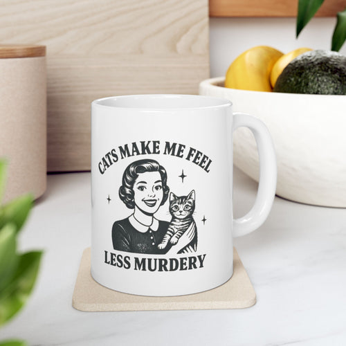 White mug with a graphic of a woman holding a cat and text 'CATS MAKE ME FEEL LESS MURDERY' on a wooden surface.
