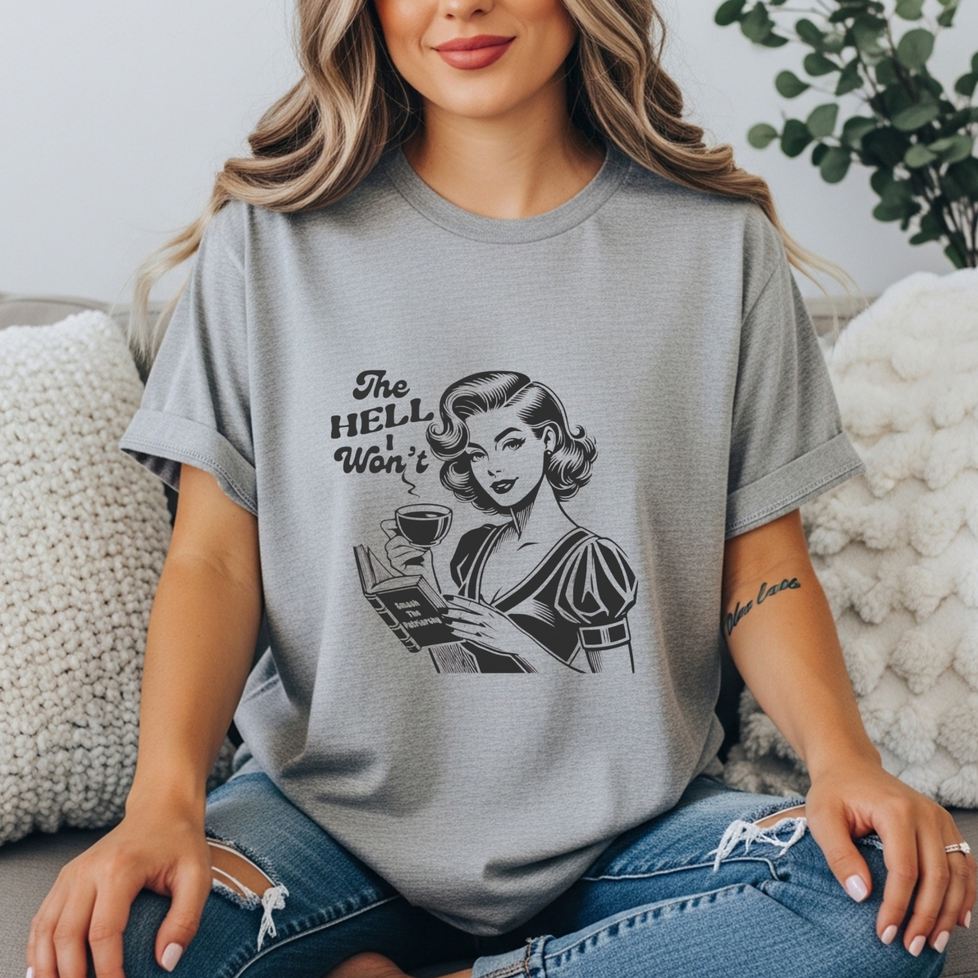 Lifestyle photo of a woman in a heather gray  feminist graphic tee. Vintage retro illustration of a 1950s woman with coffee and a book that reads “Smash the Patriarchy.” Shirt text says “The Hell I Won’t.” Funny sarcastic women’s shirt, feminist gift, retro graphic tee, bold statement shirt for women.