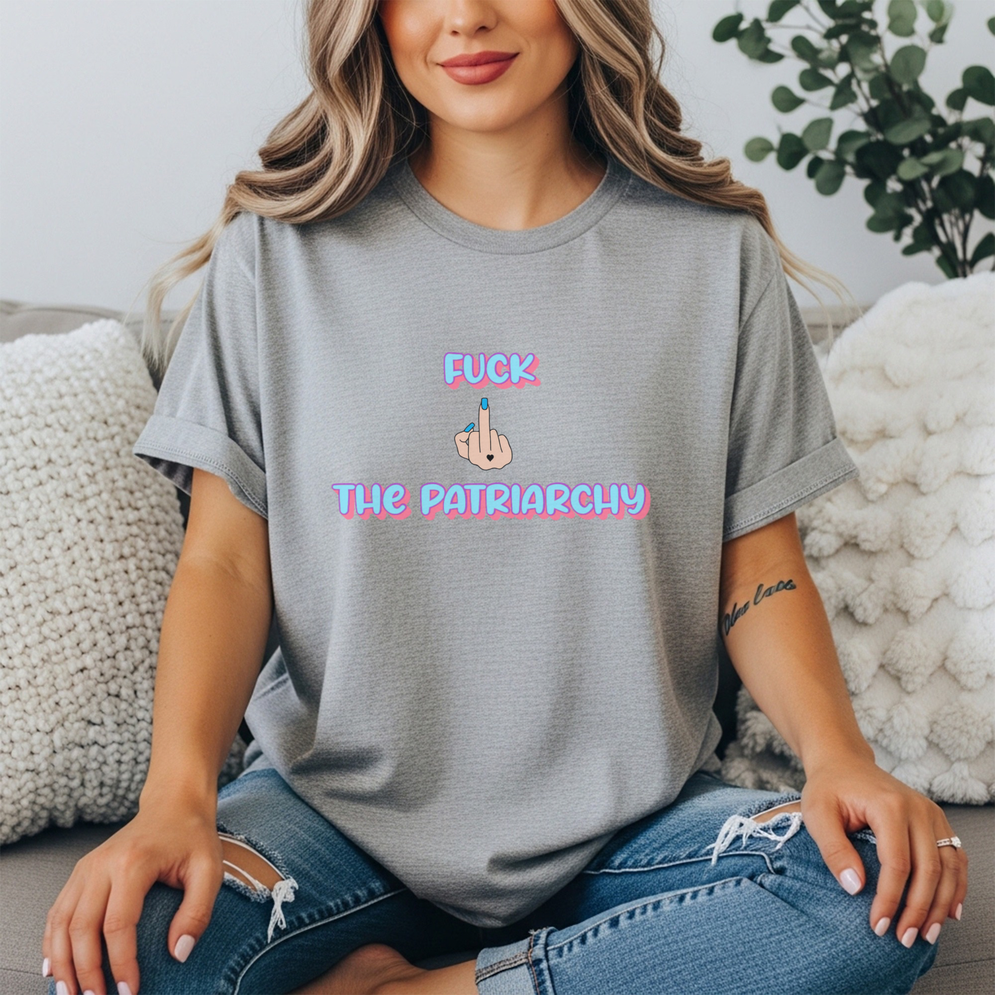 Women’s Gray  tee with “F*ck The Patriarchy” printed in pink and blue gradient bubble letters with an illustrated middle finger graphic between the words, modeled by a woman seated casually in jeans.